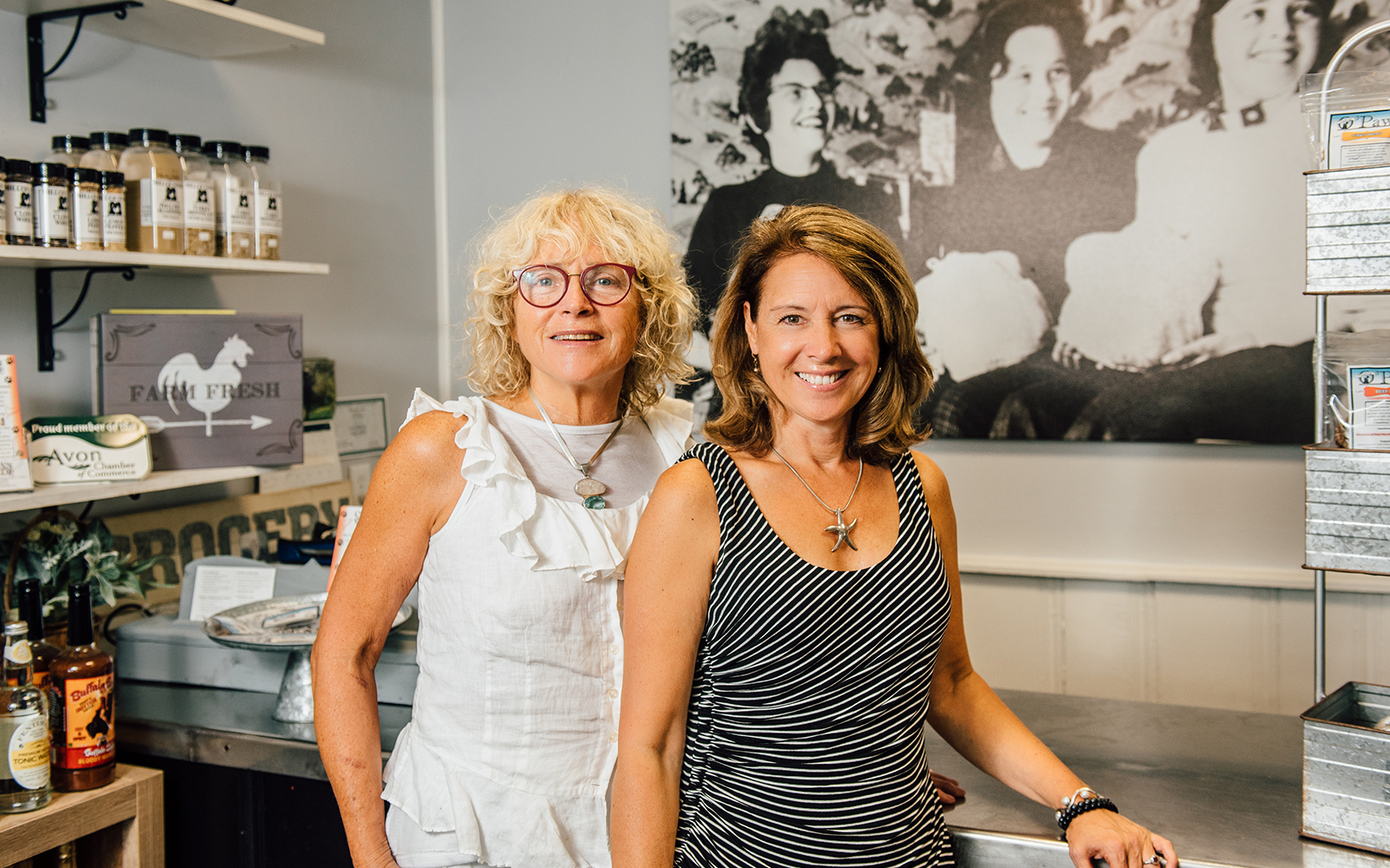 Cal Miller-Stevens, left, and her niece Capri Frank pose for a photo inside the store at Miller Foods, Inc., a fourth-generation, family owned and operated food business located in Avon, Conn. Behind them is a photo taken in the early 1960s, in the same location. From left is family matriarch Margaret "Oma" Miller and her two daughters, Sandi Trudeau (Frank's mother) and Miller-Stevens. (Nathan Oldham / UConn School of Business)