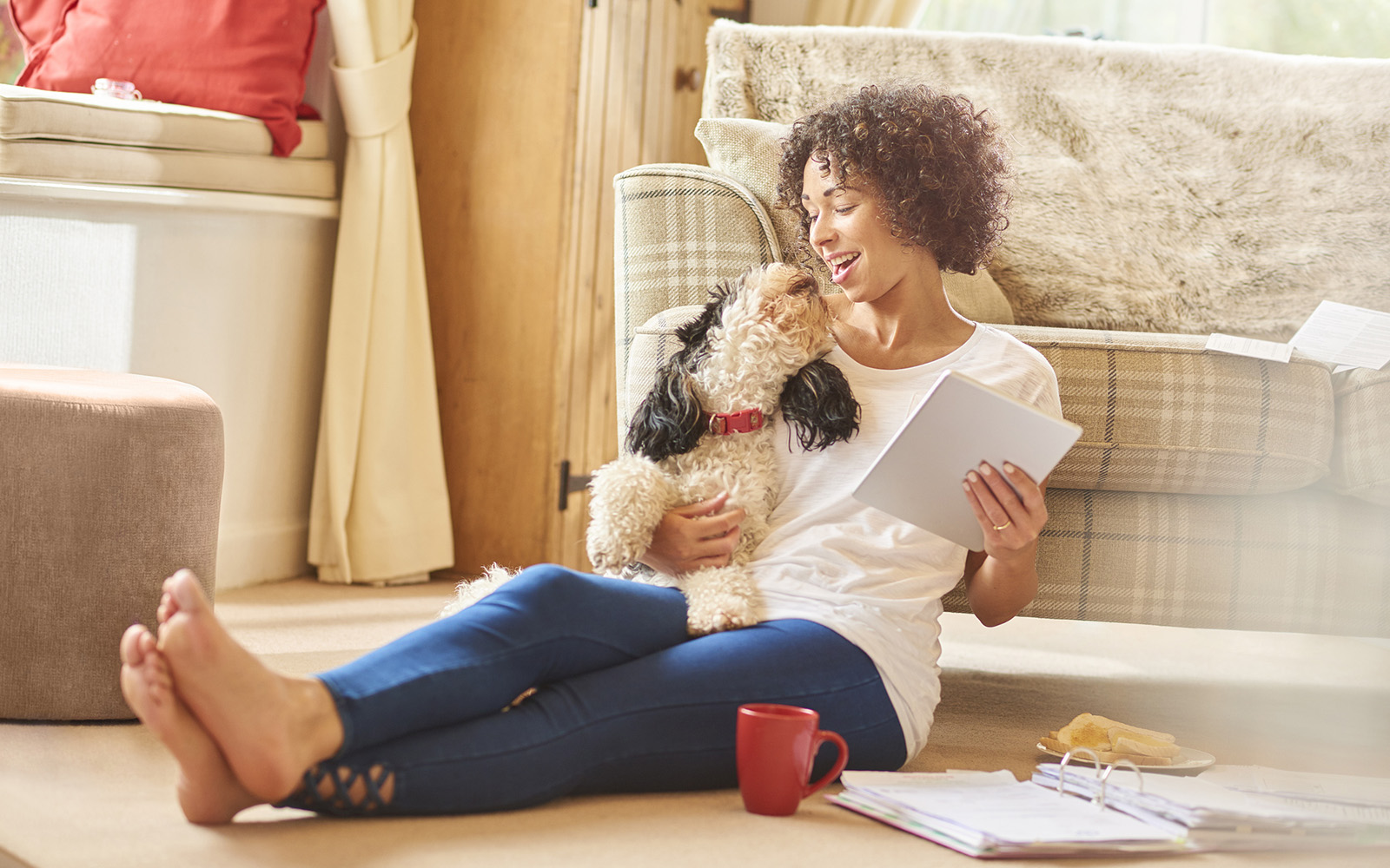 a woman sits in her lounge and browses on her digital tablet whilst sorting her paperwork with her dog