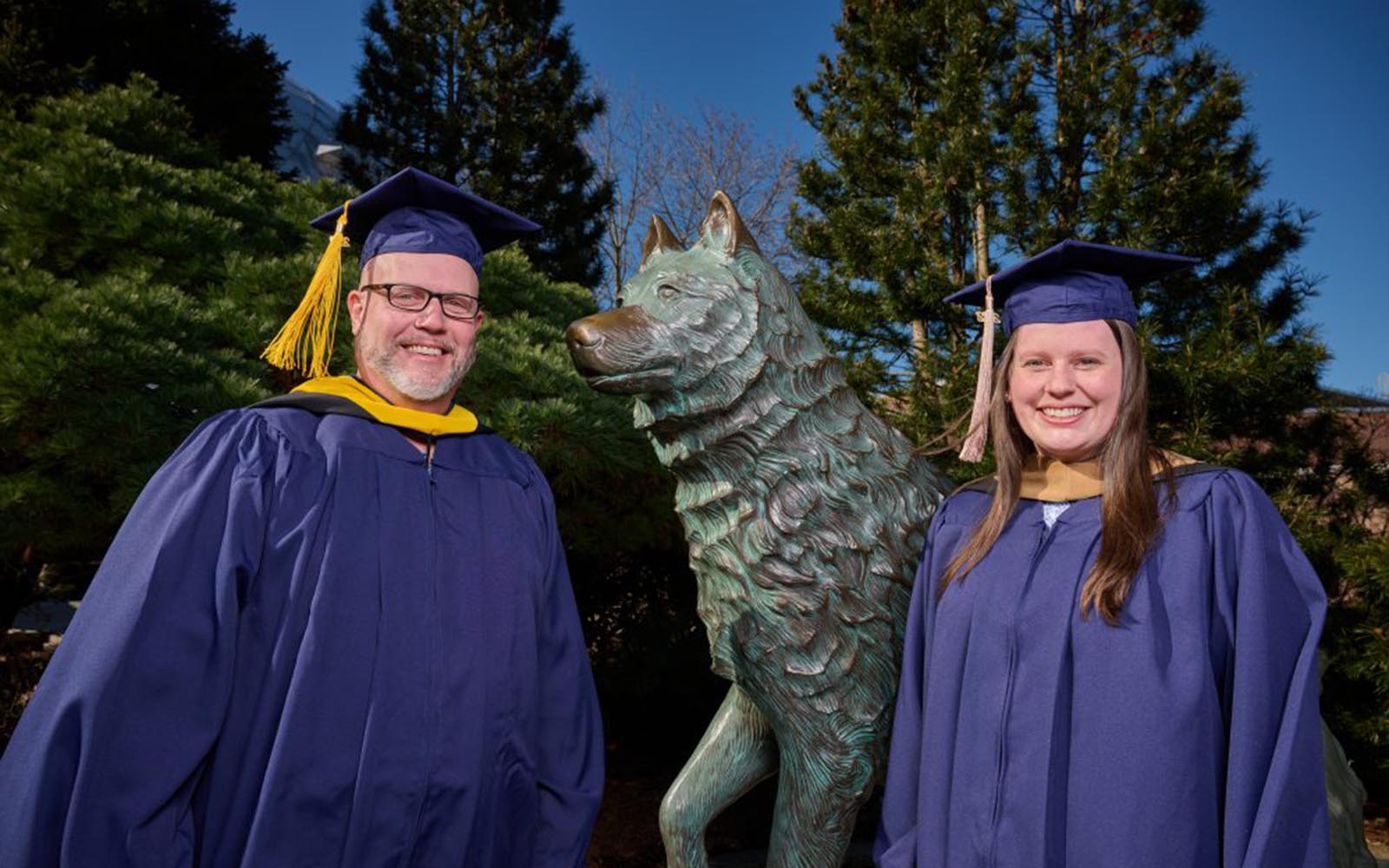 Kyle Muncy, director of brand partnerships and trademark management and his daughter Taylor Muncy, wear their caps and gowns while standing next to the Jonathan the Husky statue.