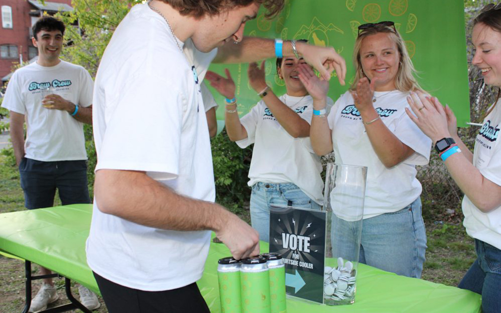 A satisfied customer votes for Courtside Cooler by dropping a bottle cap into a container, while some of the students who made the beer applaud (Claire Tremont / UConn College of Engineering)