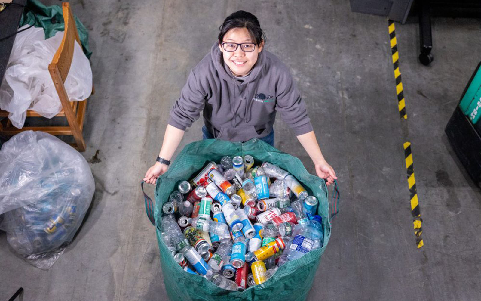 A student in a hoodie with a "Project Raccoon" logo poses with a large bin of recyclable plastic bottles