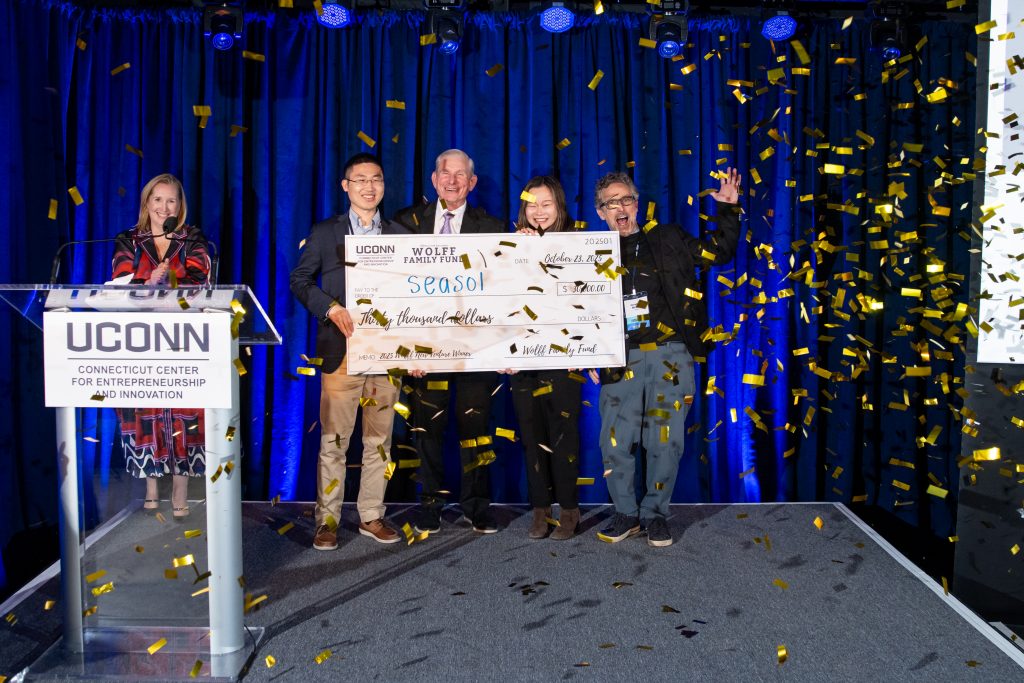 A woman stands behind a white podium while a group behind her holds a large novelty check as confetti falls.