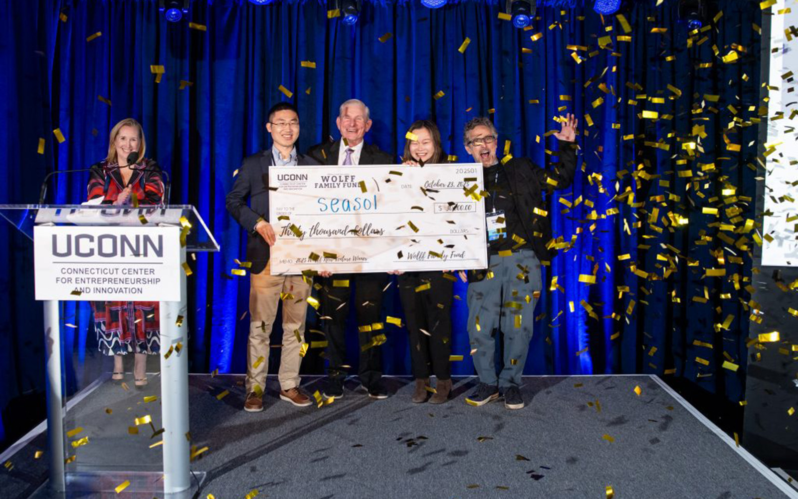 A woman stands behind a white podium while a group behind her holds a large novelty check as confetti falls.