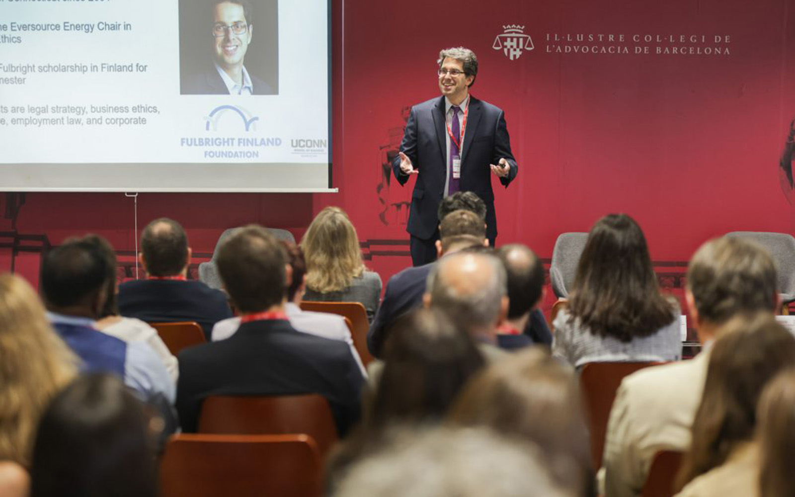A man in a suit standing before a red background addresses a large audience.