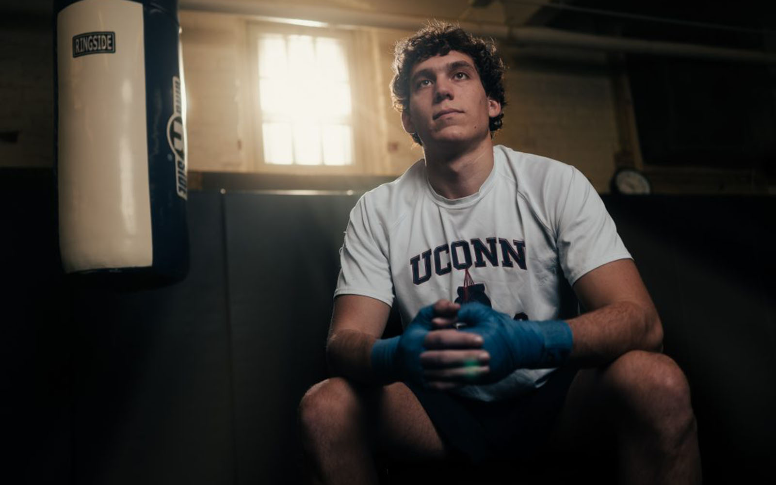 A young man in a UConn t-shirt sits next to a heavy punching bag.