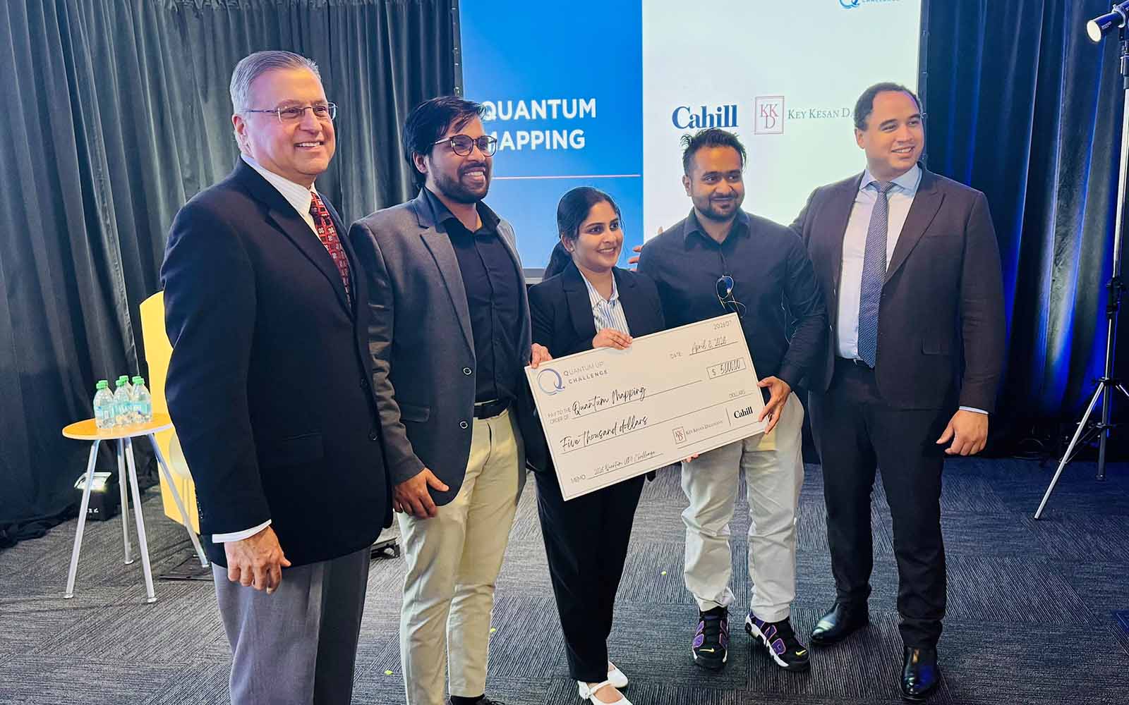 Five people pose for a group photo, with three of them holding a large novelty check.