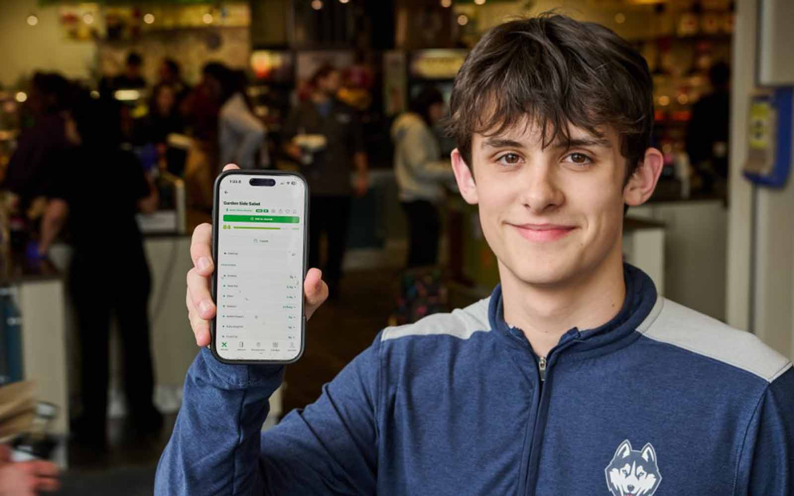 A young man with brown hair holds up a a phone to show an app on the screen
