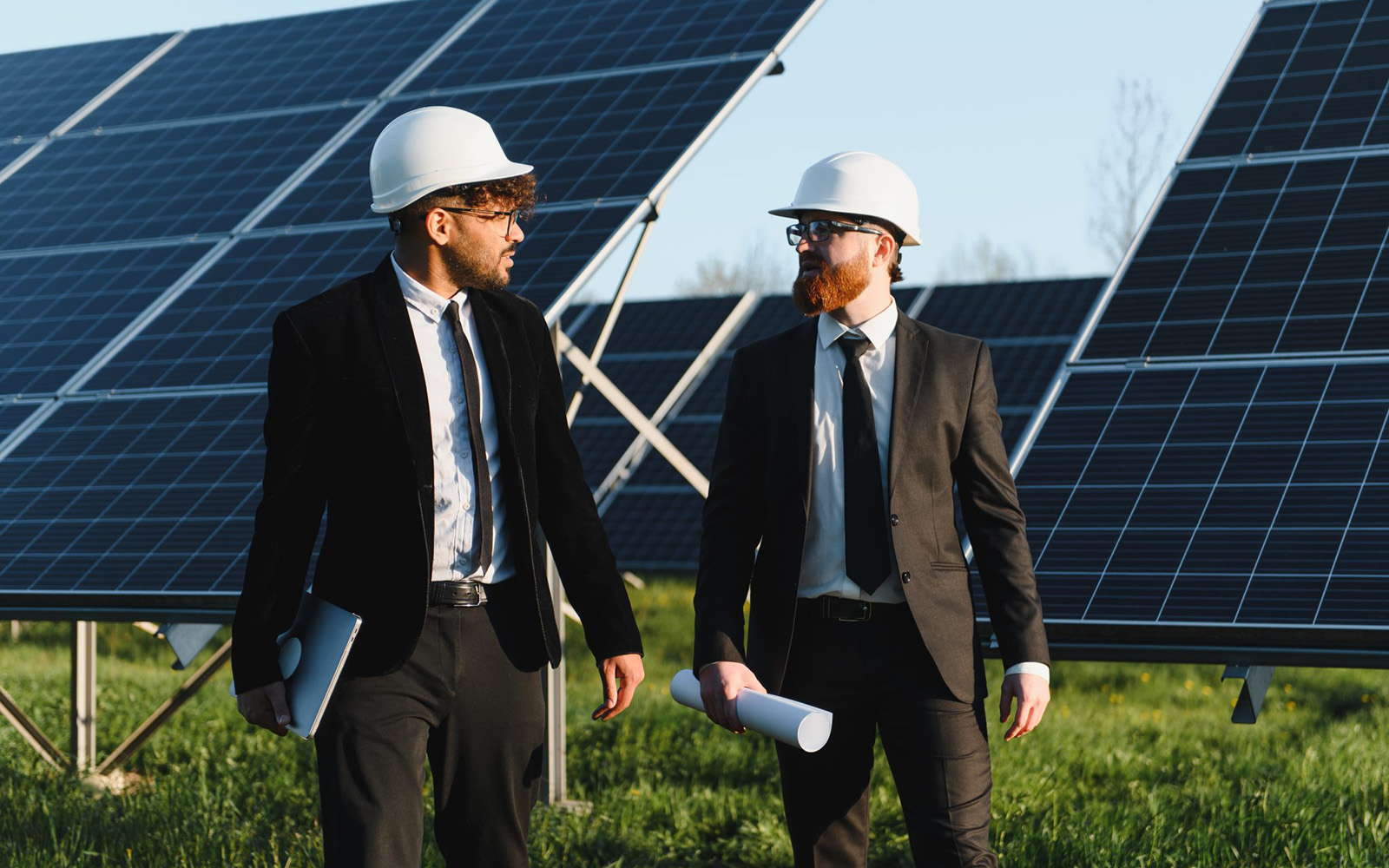 Two businessmen wearing suits and hard hats walking and discussing next to solar panels in a large solar farm.