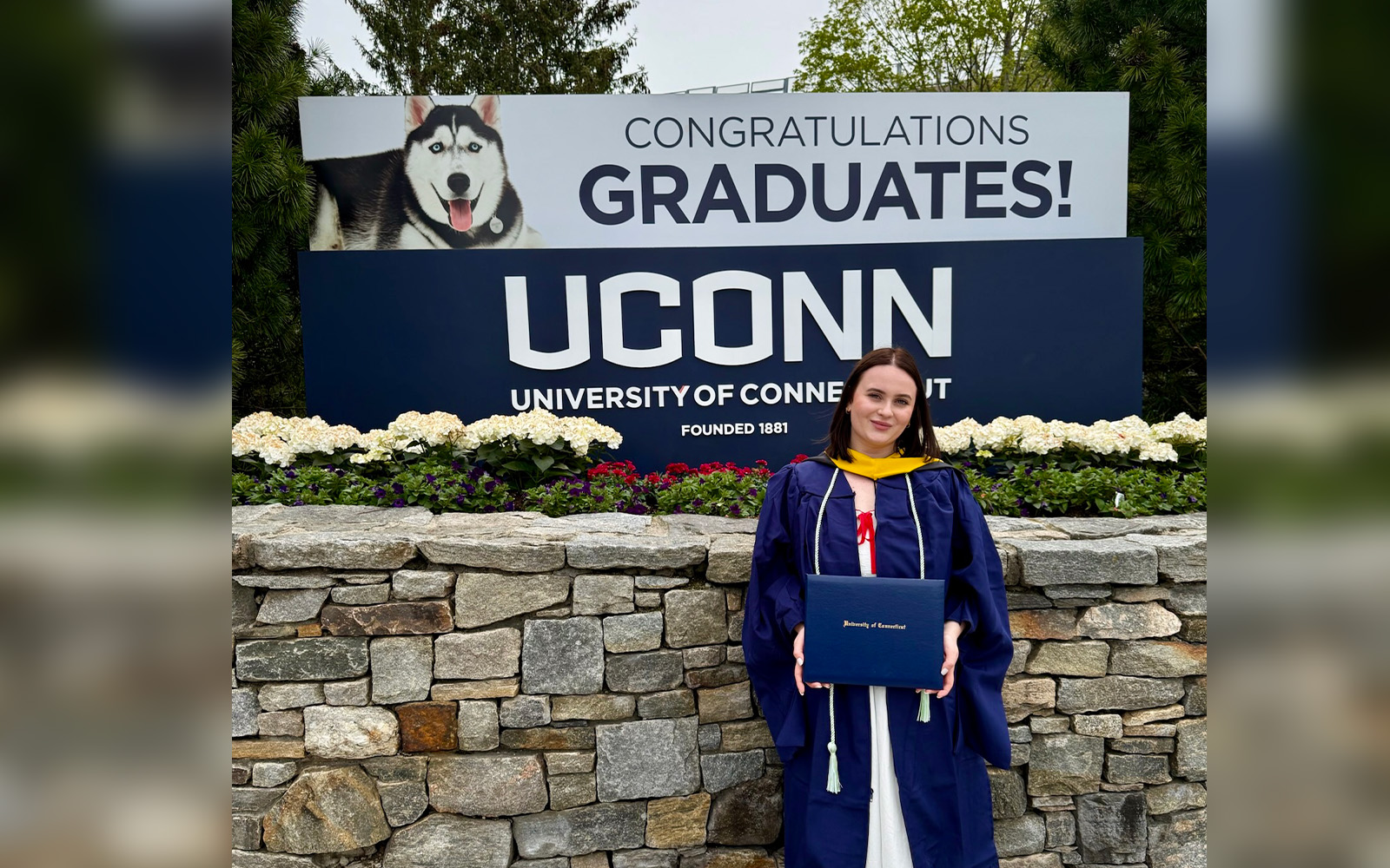 A woman in graduation robes stands in front of the UConn sign in Storrs, CT, holding a diploma