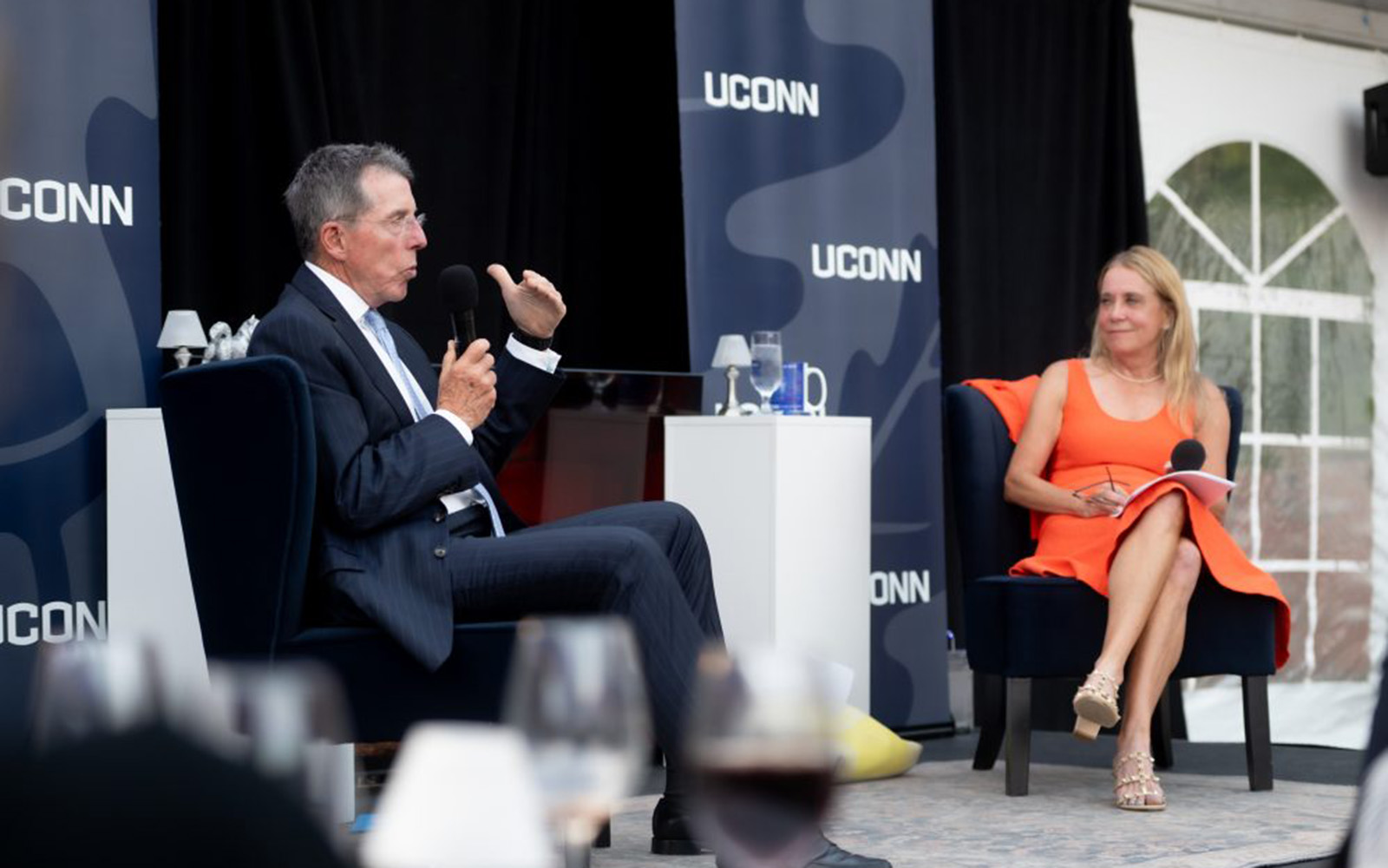 A man in a suit and a woman in an orange dress sit on stage with a small table between them and a UConn-branded backdrop behind them.