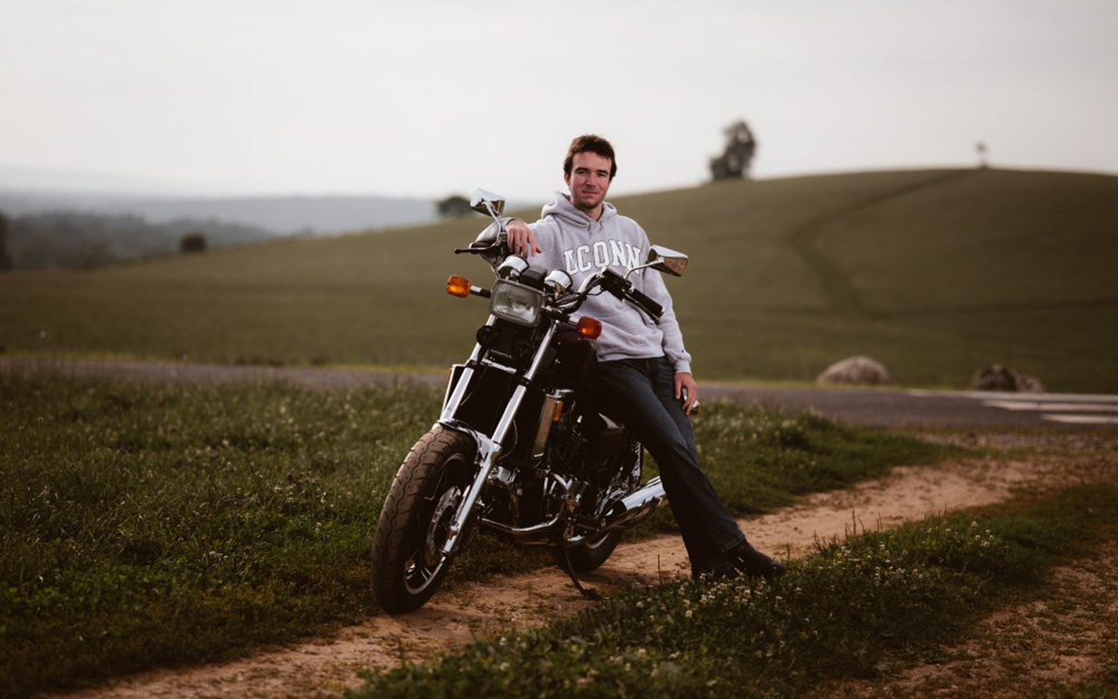 A young man in a gray sweatshirt leans on a motorcycle on Horsebarn Hill on an overcast day.
