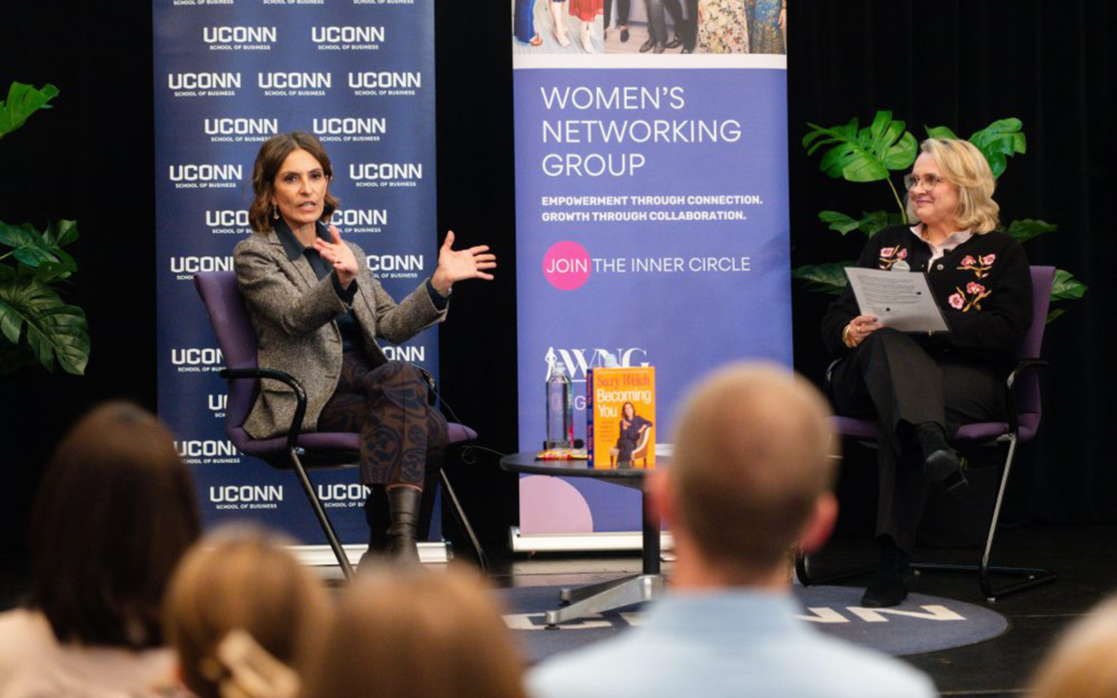 A woman in a gray blazer speaks to a crowd while seated on stage next to a woman in black. Behind them is a backdrop with UConn logos.
