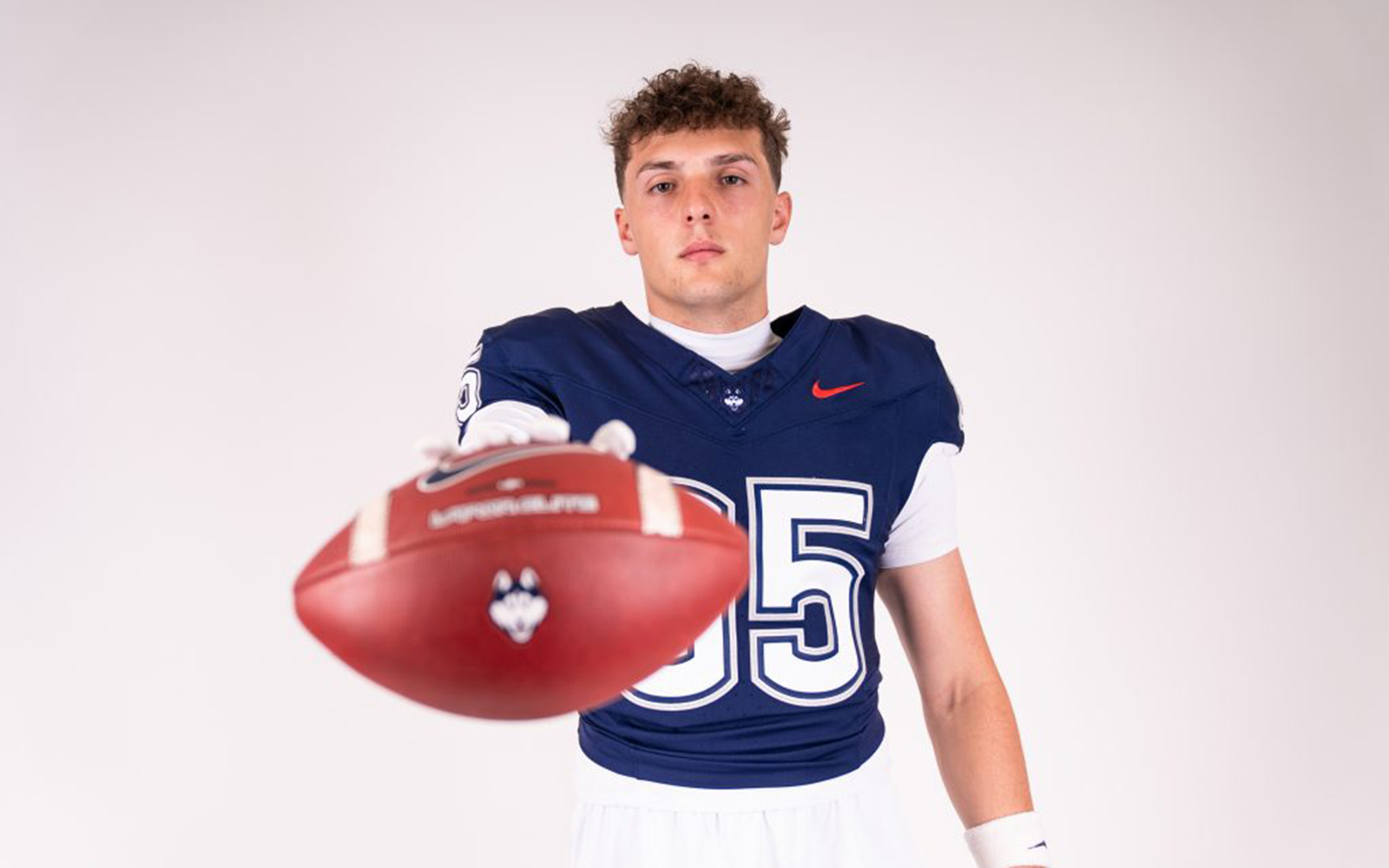A young man in a blue football jersey holds a football toward the photographer.